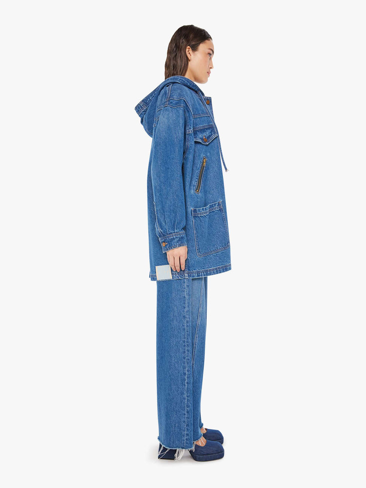 Side view of a woman oversized denim jacket with a drawstring hood, extra-wide panels at the shoulders, patch and zip pockets and an extra-long hem in a mid blue wash.