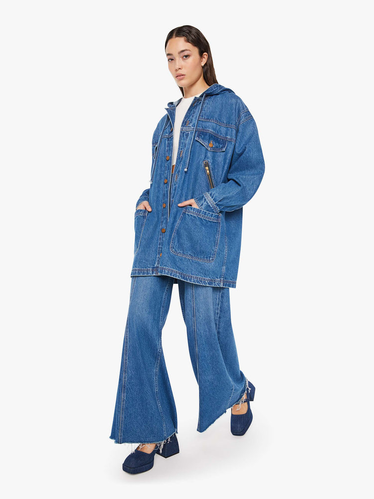 Walking view of a woman oversized denim jacket with a drawstring hood, extra-wide panels at the shoulders, patch and zip pockets and an extra-long hem in a mid blue wash.