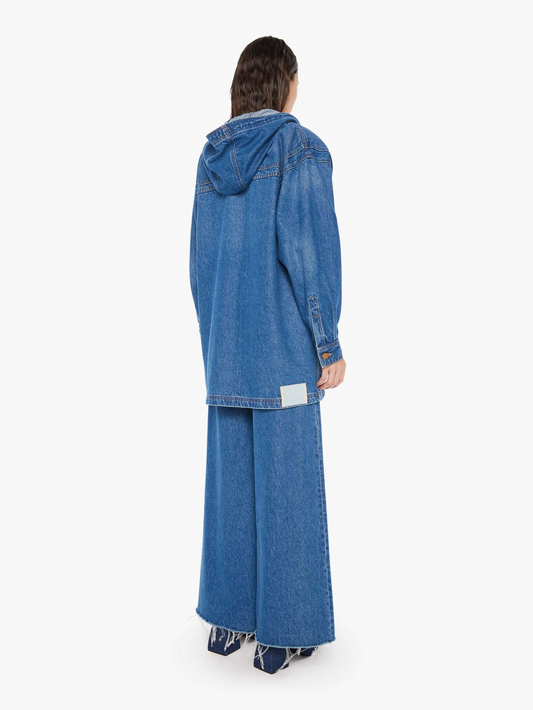 Back view of a woman oversized denim jacket with a drawstring hood, extra-wide panels at the shoulders, patch and zip pockets and an extra-long hem in a mid blue wash.