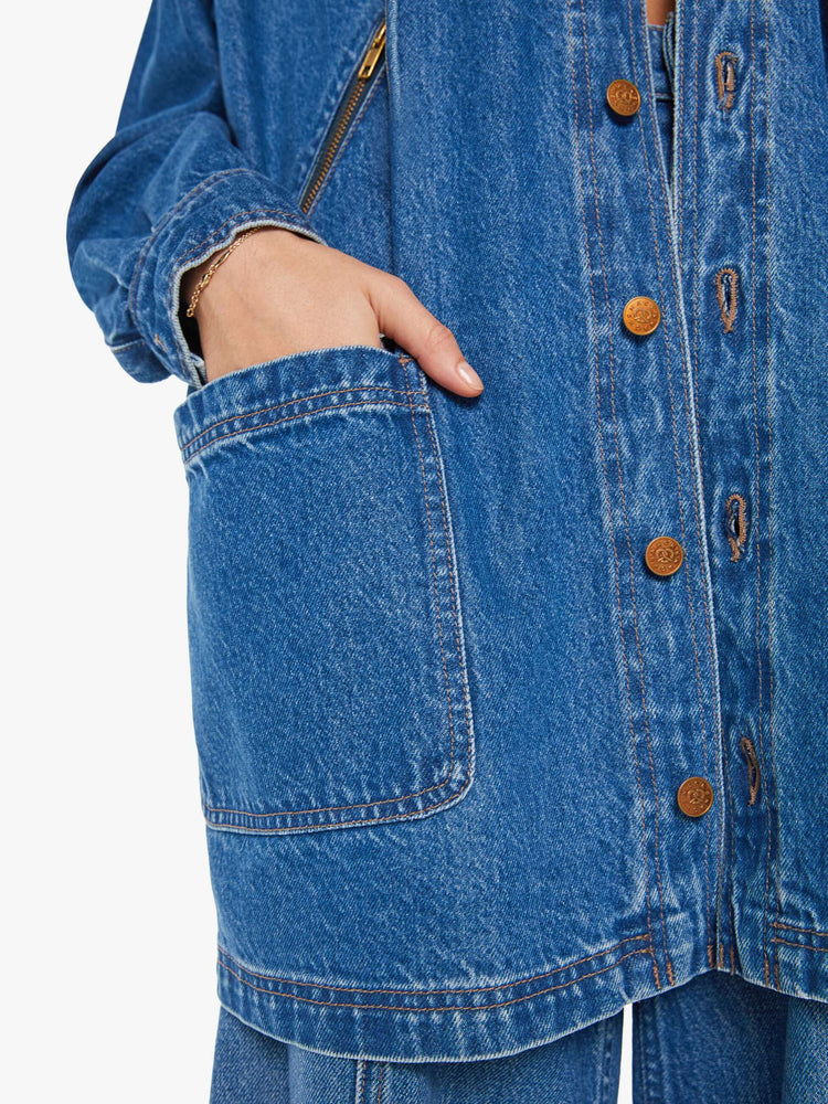 Pocket close up view of a woman oversized denim jacket with a drawstring hood, extra-wide panels at the shoulders, patch and zip pockets and an extra-long hem in a mid blue wash.