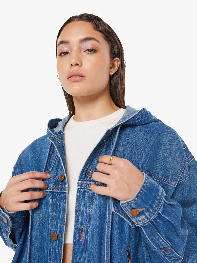 Close up view of a woman oversized denim jacket with a drawstring hood, extra-wide panels at the shoulders, patch and zip pockets and an extra-long hem in a mid blue wash.
