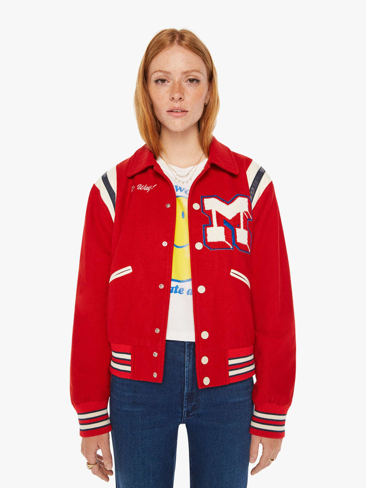 Front view of a woman red vintage-inspired varsity jacket with drop shoulders, slit pockets, ribbed hems and snaps down the front with faux leather stripes on the shoulders and a M shaped patch.