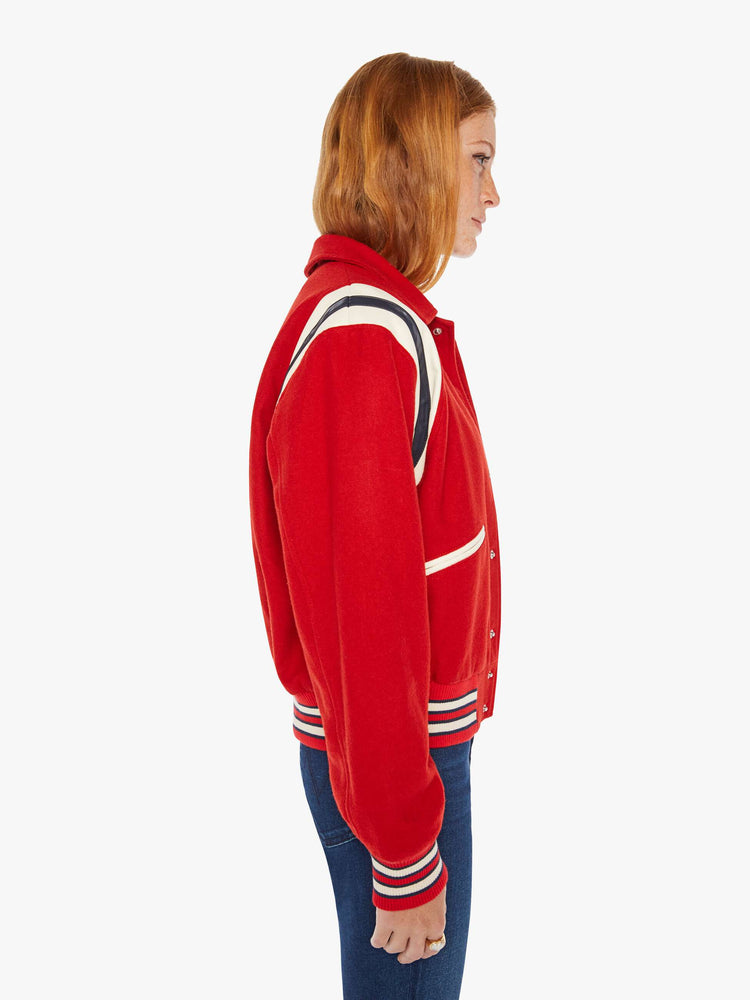 Side view of a woman red vintage-inspired varsity jacket with drop shoulders, slit pockets, ribbed hems and snaps down the front with faux leather stripes on the shoulders and a M shaped patch.