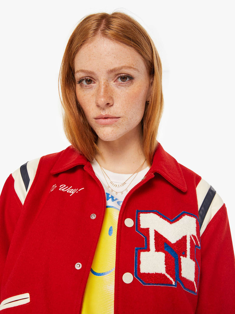 Close up view of a woman red vintage-inspired varsity jacket with drop shoulders, slit pockets, ribbed hems and snaps down the front with faux leather stripes on the shoulders and a M shaped patch.
