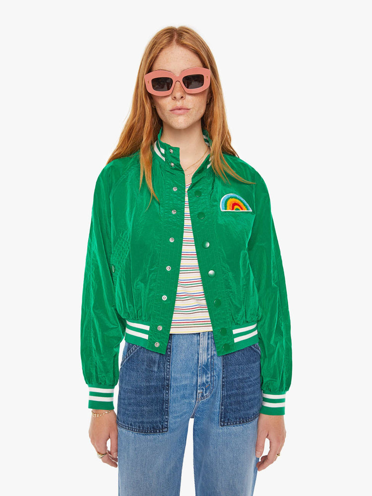 Front view of a woman green lightweight windbreaker with a high neck, bat-wing sleeves, slit pockets, ribbed hems and a cropped fit with white stripes at the hem and a rainbow patch.