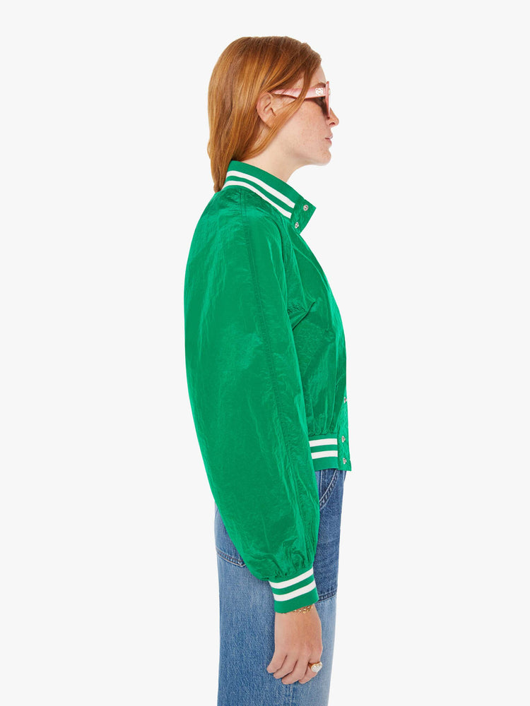 Side view of a woman green lightweight windbreaker with a high neck, bat-wing sleeves, slit pockets, ribbed hems and a cropped fit with white stripes at the hem and a rainbow patch.