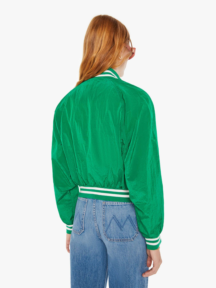 Back view of a woman green lightweight windbreaker with a high neck, bat-wing sleeves, slit pockets, ribbed hems and a cropped fit with white stripes at the hem and a rainbow patch.