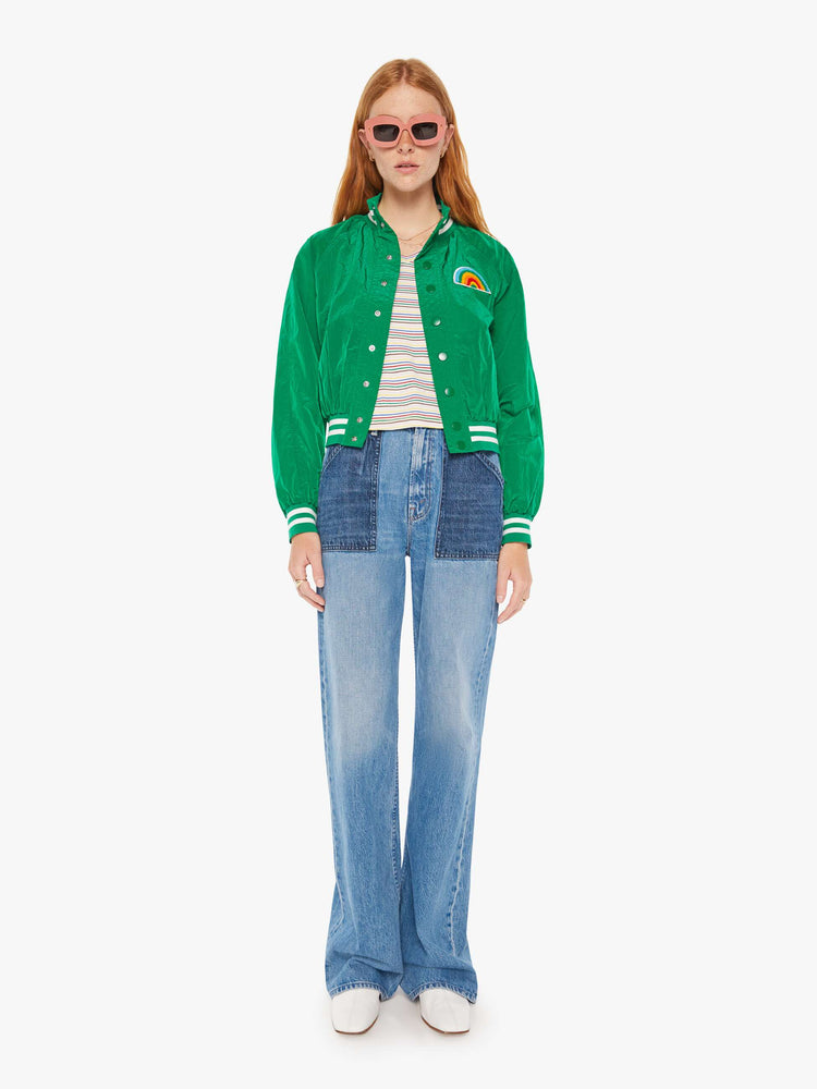Full body view of a woman green lightweight windbreaker with a high neck, bat-wing sleeves, slit pockets, ribbed hems and a cropped fit with white stripes at the hem and a rainbow patch.