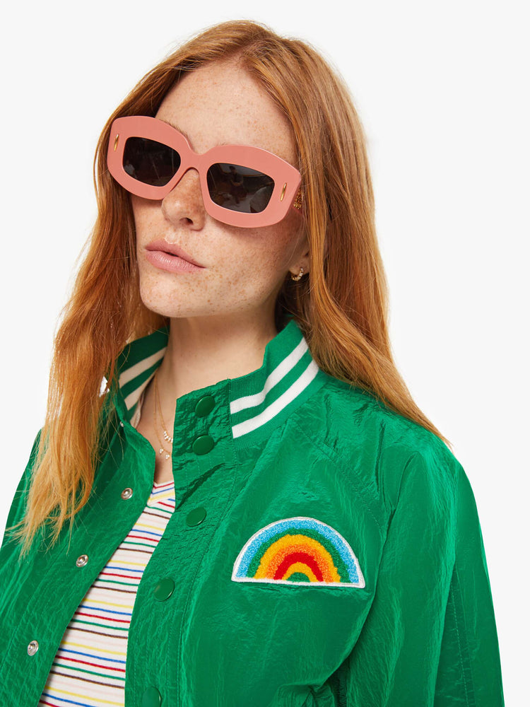 Close up view of a woman green lightweight windbreaker with a high neck, bat-wing sleeves, slit pockets, ribbed hems and a cropped fit with white stripes at the hem and a rainbow patch.