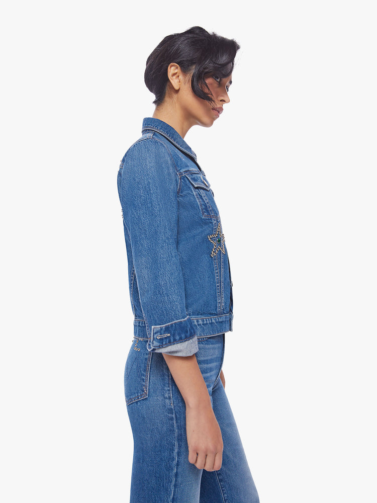 Side view of a woman classic denim jacket with front patch pockets and slightly cropped hems in a med blue wash with studded stars, moons and text on the back.