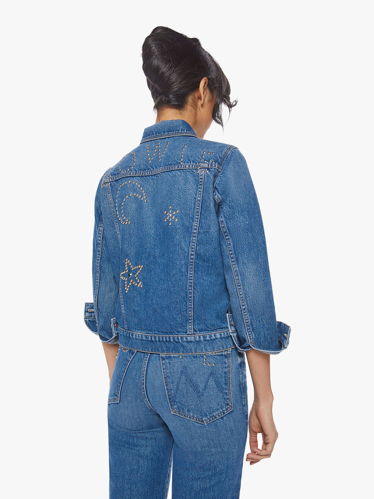Back view of a woman classic denim jacket with front patch pockets and slightly cropped hems in a med blue wash with studded stars, moons and text on the back.