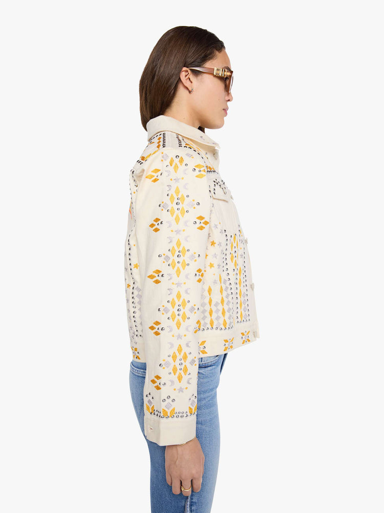 Side view of a woman in a warm cream with silver and golden-yellow embroidery, decorative rivets and oversized patches on the back, classic button-up jacket with front patch pockets and a slightly cropped fit.