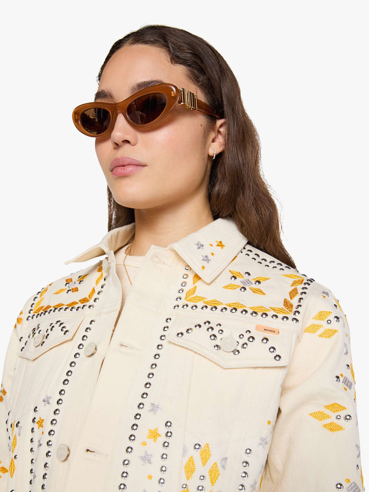 Close up view of a woman in a warm cream with silver and golden-yellow embroidery, decorative rivets and oversized patches on the back, classic button-up jacket with front patch pockets and a slightly cropped fit.