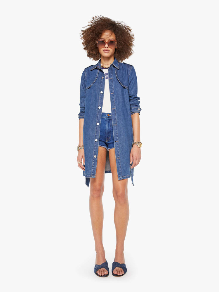 Front view of a woman in a med blue denim trench coat features a tied waist, back storm flap and a thigh-length hem. 