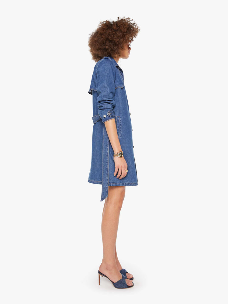 Side view of a woman in a med blue denim trench coat features a tied waist, back storm flap and a thigh-length hem. 