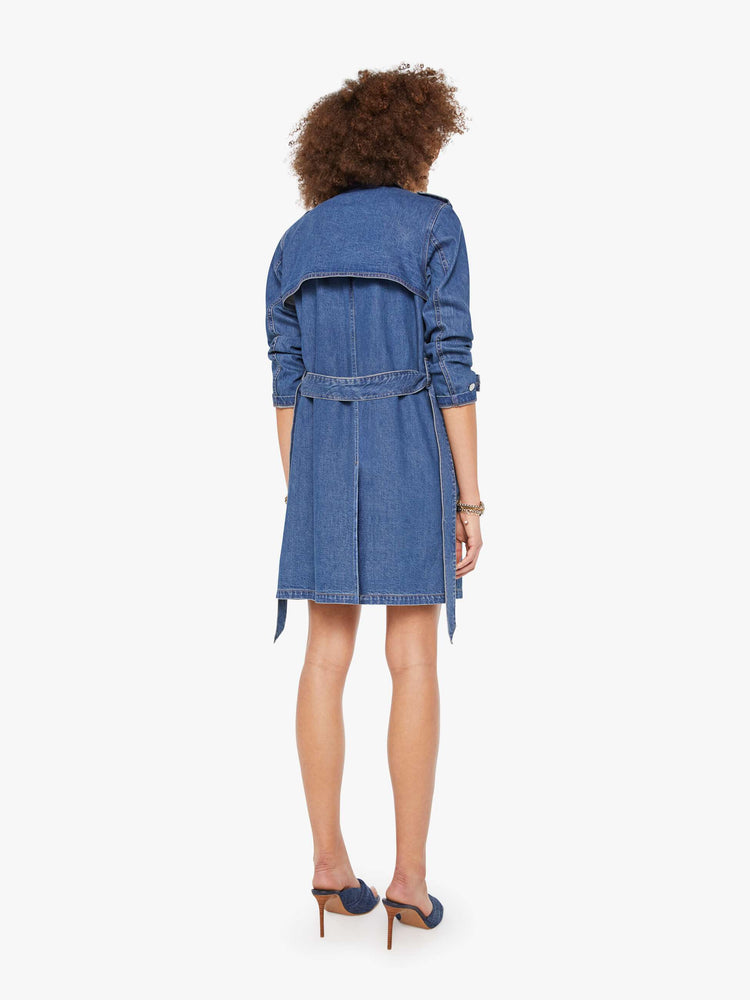 Back view of a woman in a med blue denim trench coat features a tied waist, back storm flap and a thigh-length hem. 