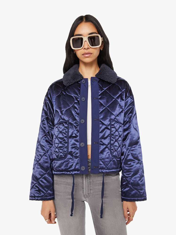 Front view of a woman in quilted collared jacket in an iridescent dark blue with a faux-sherpa collar and tonal buttons.