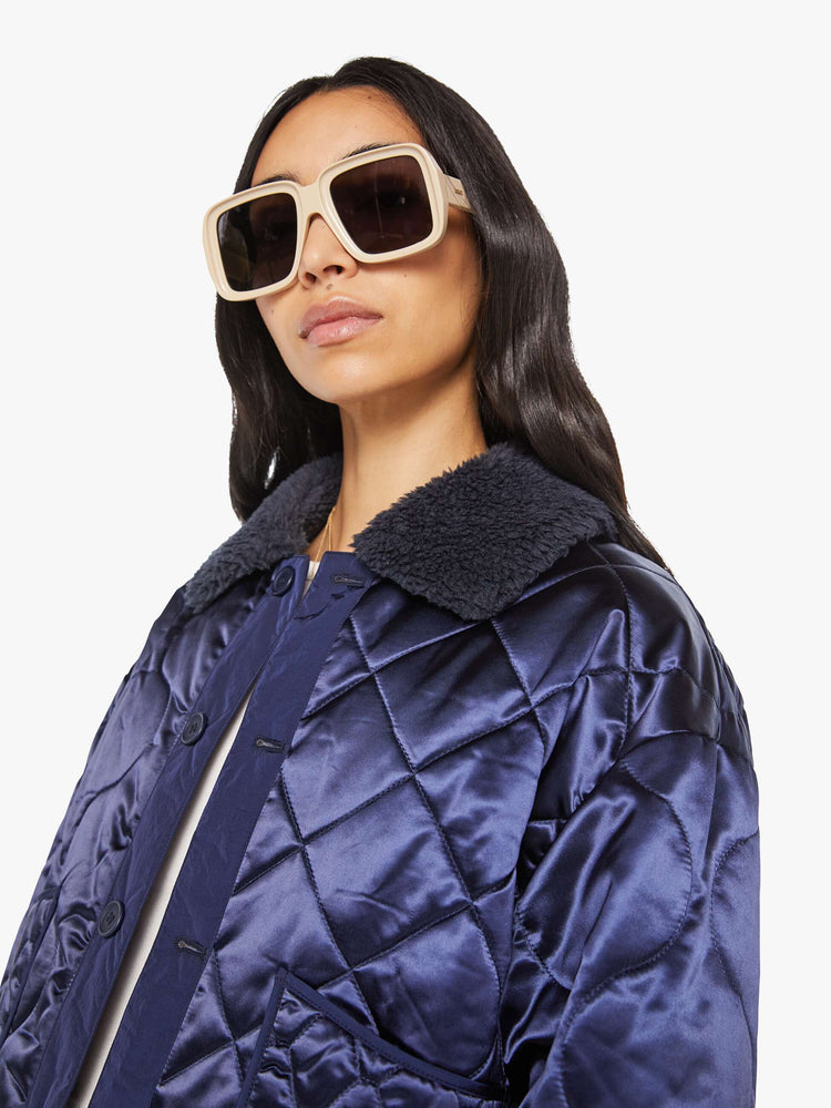 Close up view of a woman in quilted collared jacket in an iridescent dark blue with a faux-sherpa collar and tonal buttons.