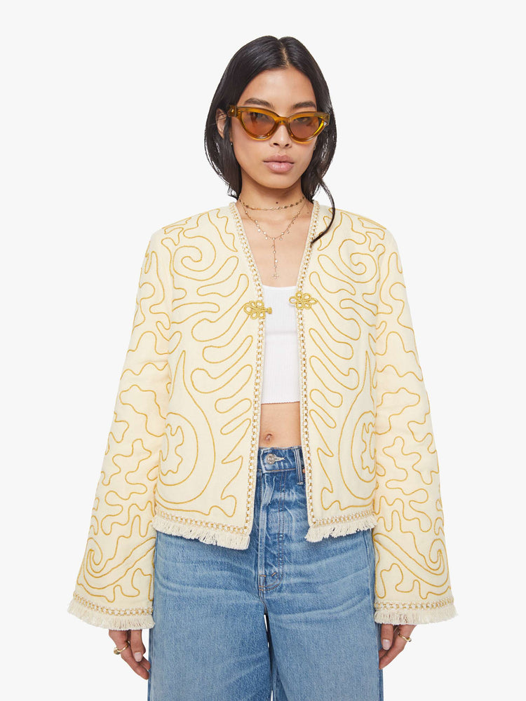 Front view of a woman in off white with gold embellishments V-neck jacket with bell sleeves, a button closure and fringed hem.