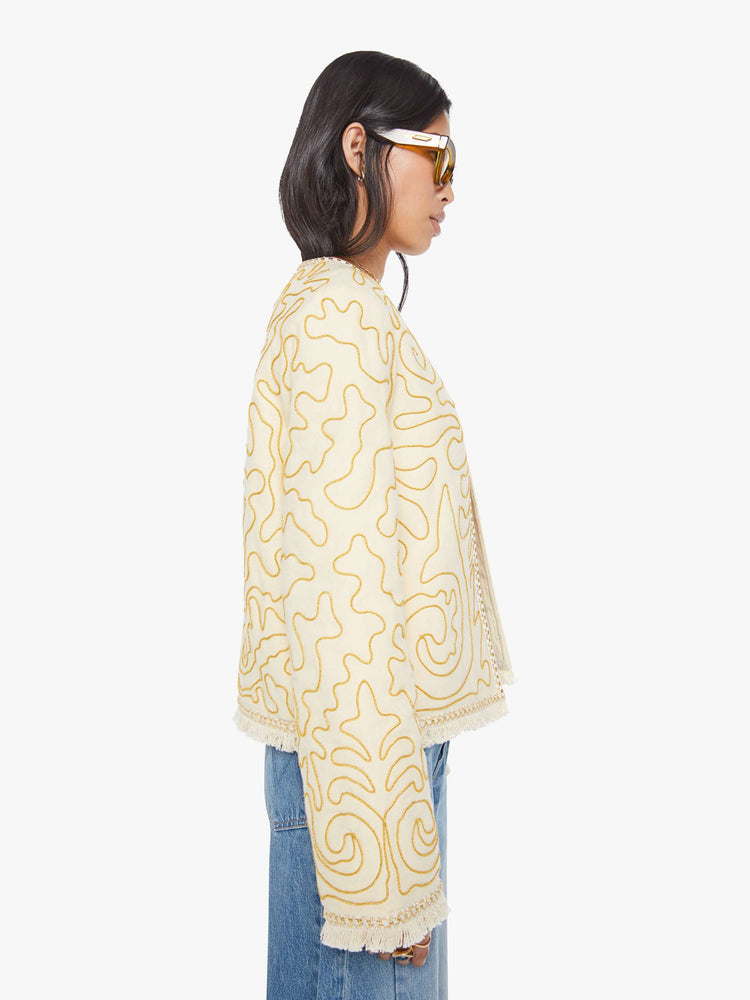Side view of a woman in off white with gold embellishments V-neck jacket with bell sleeves, a button closure and fringed hem.