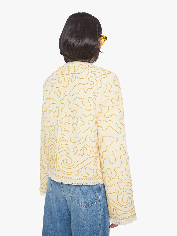 Back view of a woman in off white with gold embellishments V-neck jacket with bell sleeves, a button closure and fringed hem.