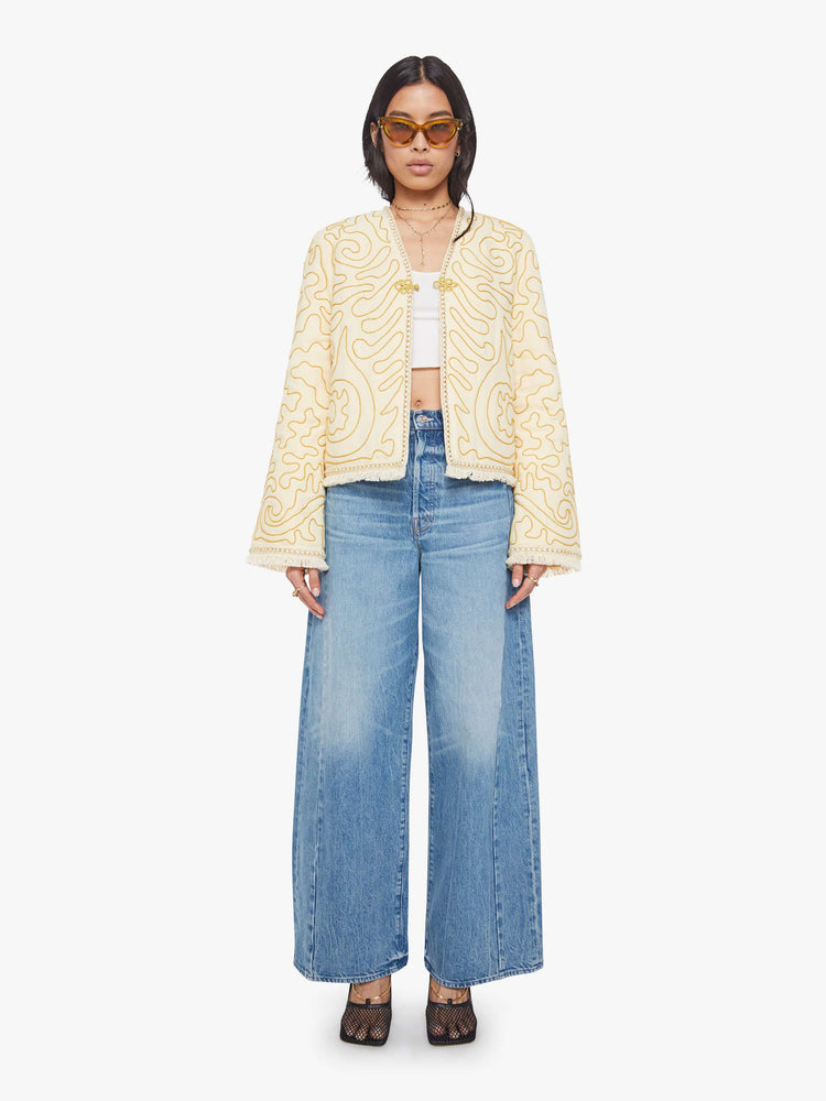 Full body view of a woman in off white with gold embellishments V-neck jacket with bell sleeves, a button closure and fringed hem.