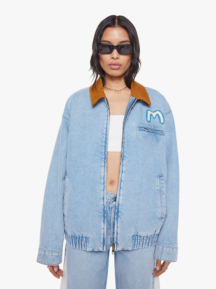 Front view of a woman in a light blue with a hand-embroidered M patch on the chest, quilted lining, a corduroy collar oversized denim jacket is designed with slit pockets, drop shoulders, an elastic hem and zip closure. 