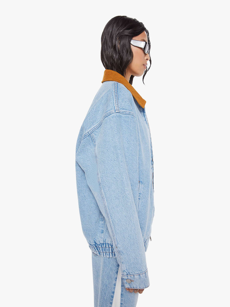 Side  view of a woman in a light blue with a hand-embroidered M patch on the chest, quilted lining, a corduroy collar oversized denim jacket is designed with slit pockets, drop shoulders, an elastic hem and zip closure. 