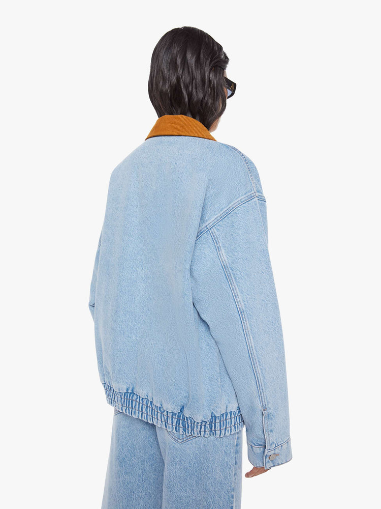 Back  view of a woman in a light blue with a hand-embroidered M patch on the chest, quilted lining, a corduroy collar oversized denim jacket is designed with slit pockets, drop shoulders, an elastic hem and zip closure. 