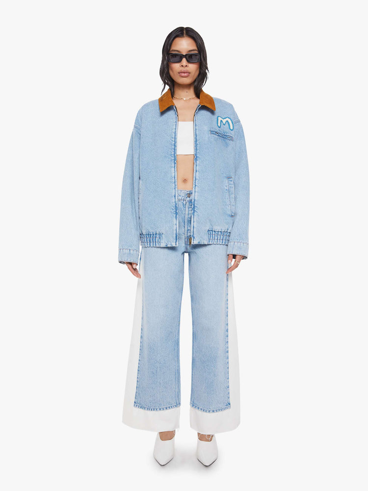 Full body  view of a woman in a light blue with a hand-embroidered M patch on the chest, quilted lining, a corduroy collar oversized denim jacket is designed with slit pockets, drop shoulders, an elastic hem and zip closure. 