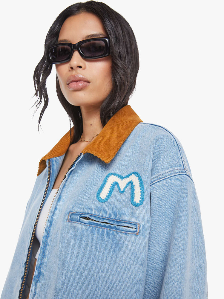 Close up  view of a woman in a light blue with a hand-embroidered M patch on the chest, quilted lining, a corduroy collar oversized denim jacket is designed with slit pockets, drop shoulders, an elastic hem and zip closure. 