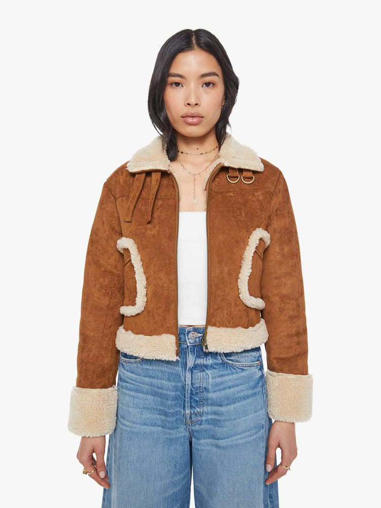 Front view of a woman in a caramel faux suede  cropped jacket with front patch pockets, buckle details on the collar and sides and a boxy fit. 