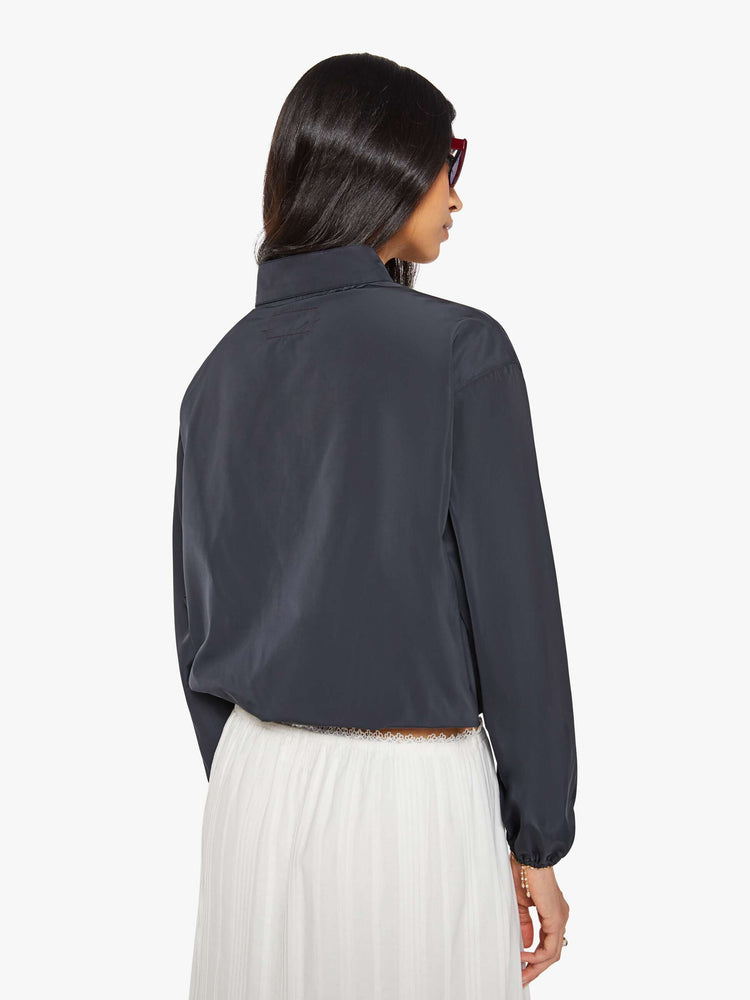 Back view of a woman in a navy with red and white stripe vintage-inspired windbreaker with a short stacked collar that buttons, a zip closure, drop shoulders, balloon sleeves, a small patch pocket and a cropped drawstring hem.