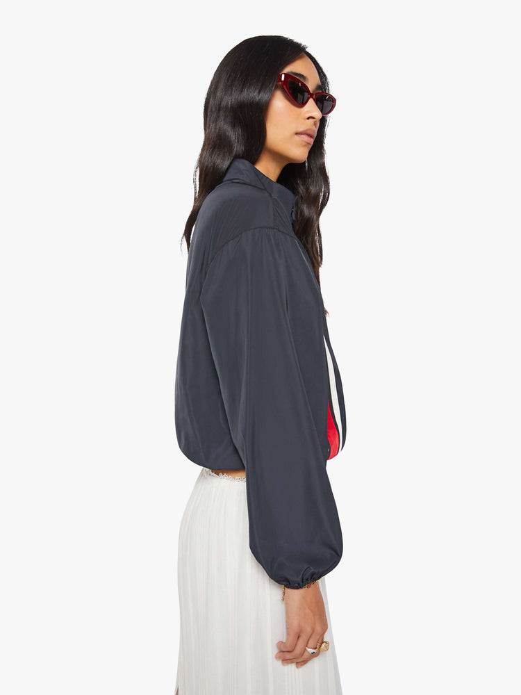 Side view of a woman in a navy with red and white stripe vintage-inspired windbreaker with a short stacked collar that buttons, a zip closure, drop shoulders, balloon sleeves, a small patch pocket and a cropped drawstring hem.