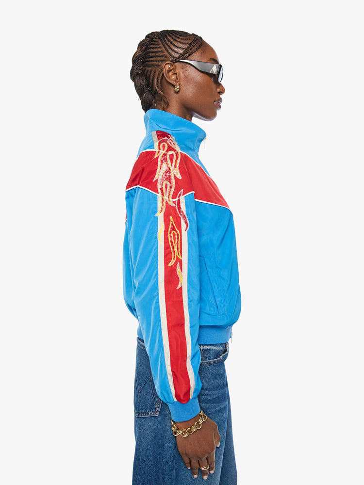 Side view of a woman in a glossy vintage blue letterman jacket with angled red panels across the chest, stripes down the sleeves, an M patch in yellow on the front and stitched details along the shoulders.