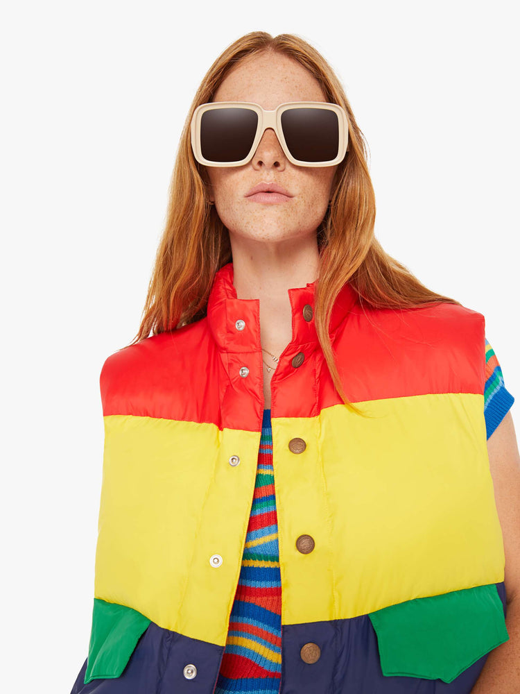 Close up view of a woman puffer vest with a funnel neck, snaps down the front and patch pockets in shades of red, yellow and navy with green details at pockets.