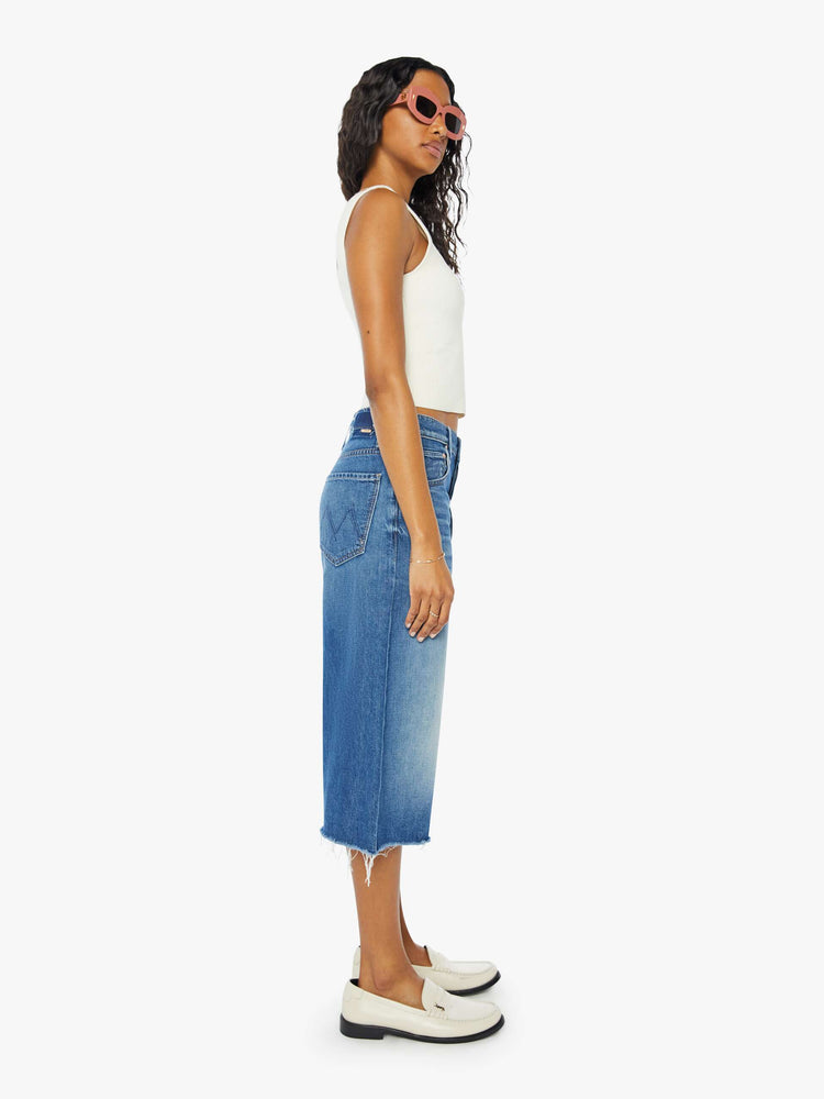 Side view of a woman wearing a medium blue wash short featuring a knee length inseam, raw hem, and wide leg, paired with a white tank top.