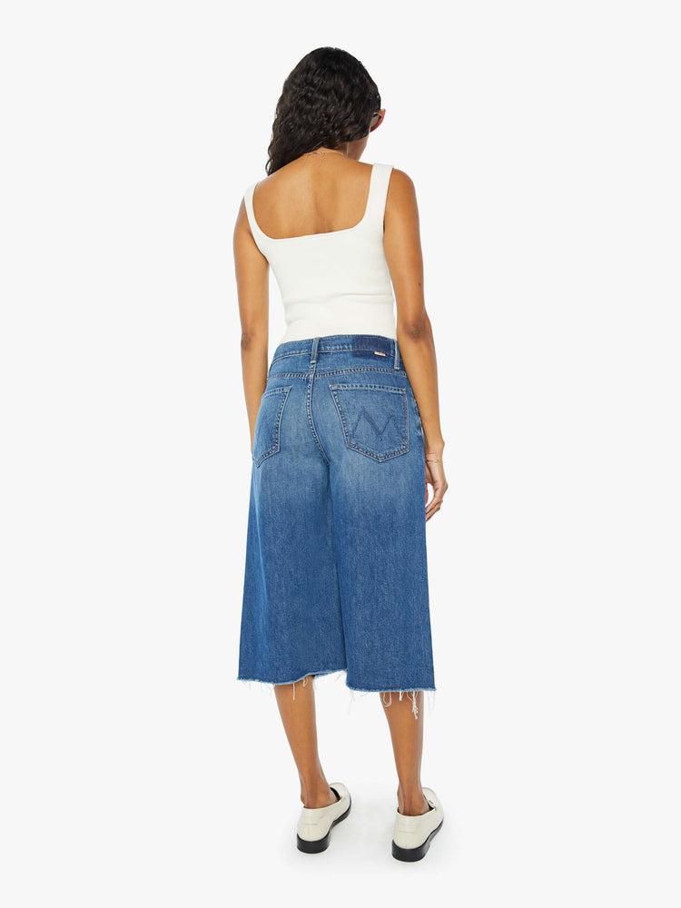 Back view of a woman wearing a medium blue wash short featuring a knee length inseam, raw hem, and wide leg, paired with a white tank top.