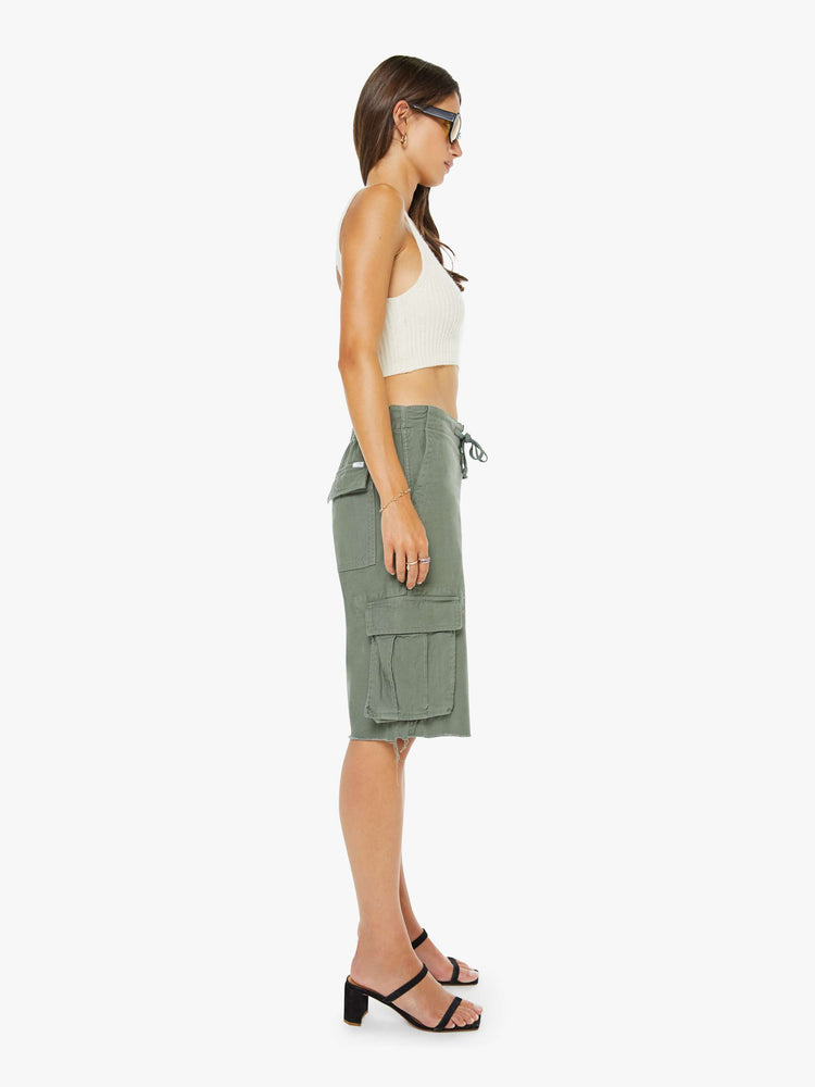 Side view of a woman in an army green shorts with a drawstring waist, zip fly, patch pockets on the thighs and a knee-length inseam with a frayed hem.