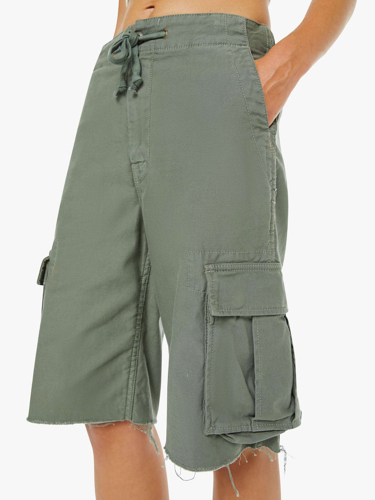 Close up view of a woman in an army green shorts with a drawstring waist, zip fly, patch pockets on the thighs and a knee-length inseam with a frayed hem.
