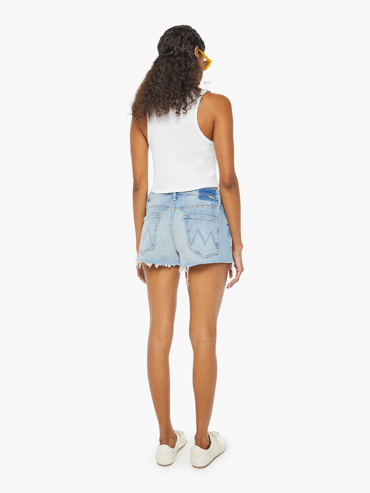 Back view of a woman in light blue denim shorts designed to sit lower on the hips with a frayed hem and whiskering, fading, and distressed details. 