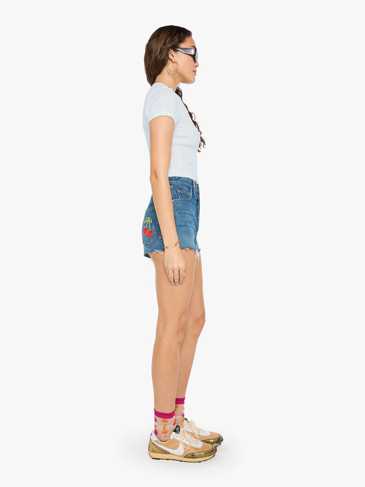 Side view of a woman in a super high-waisted denim shorts with a button fly, 2-inch inseam and a frayed hem in mid blue wash with cherries embroidered.