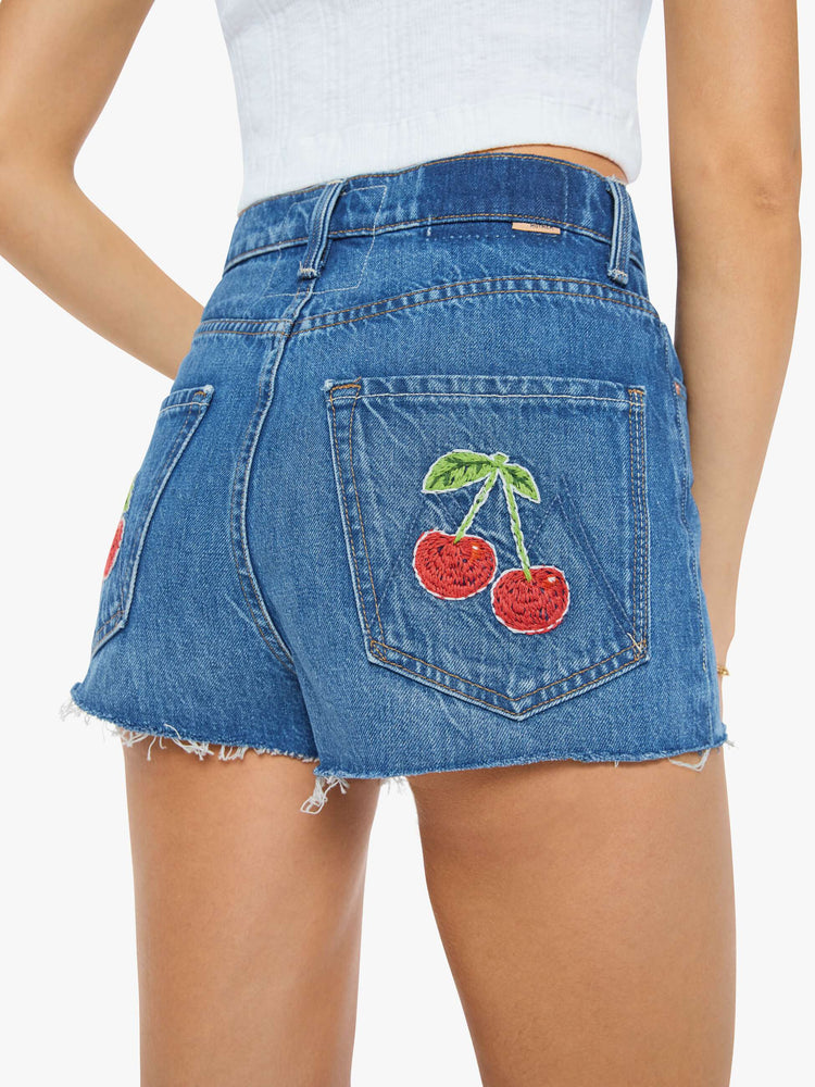 Close up view of a woman in a super high-waisted denim shorts with a button fly, 2-inch inseam and a frayed hem in mid blue wash with cherries embroidered.