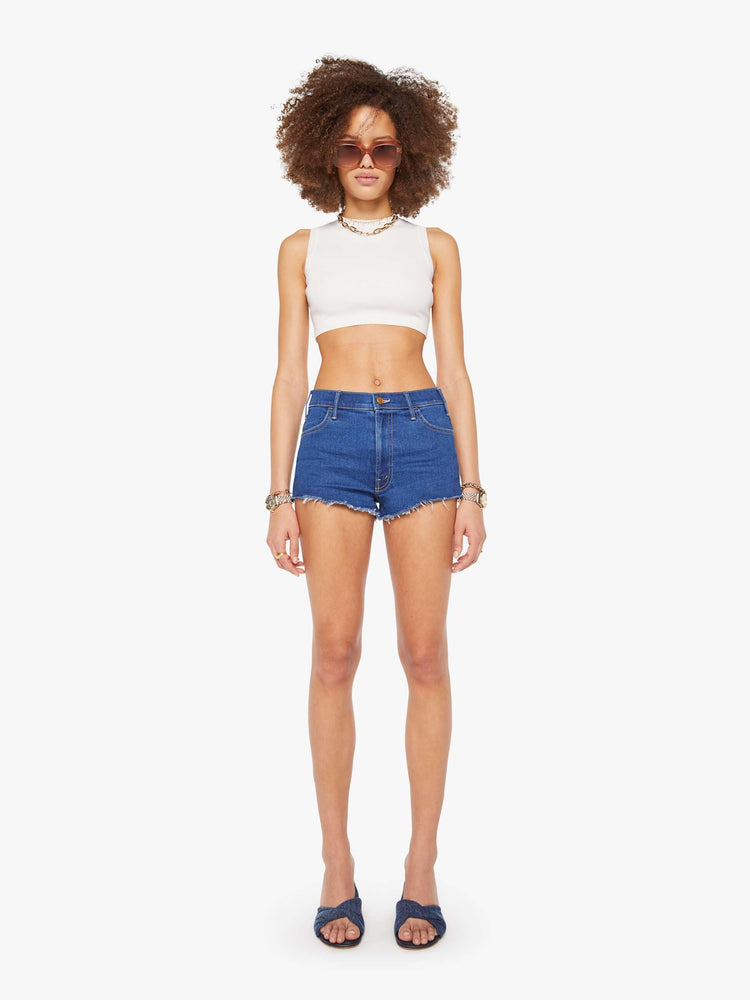 Front view of a woman in a  midblue high-waisted denim shorts with a zip fly, short inseam and frayed hem