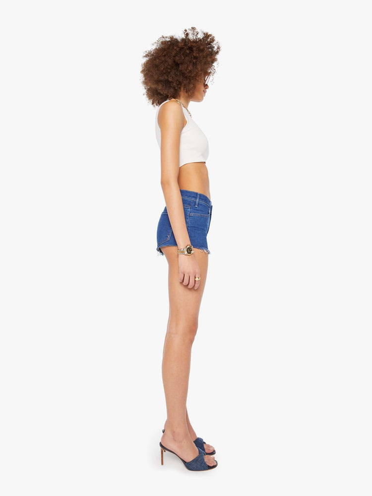 Side view of a woman in a  midblue high-waisted denim shorts with a zip fly, short inseam and frayed hem.