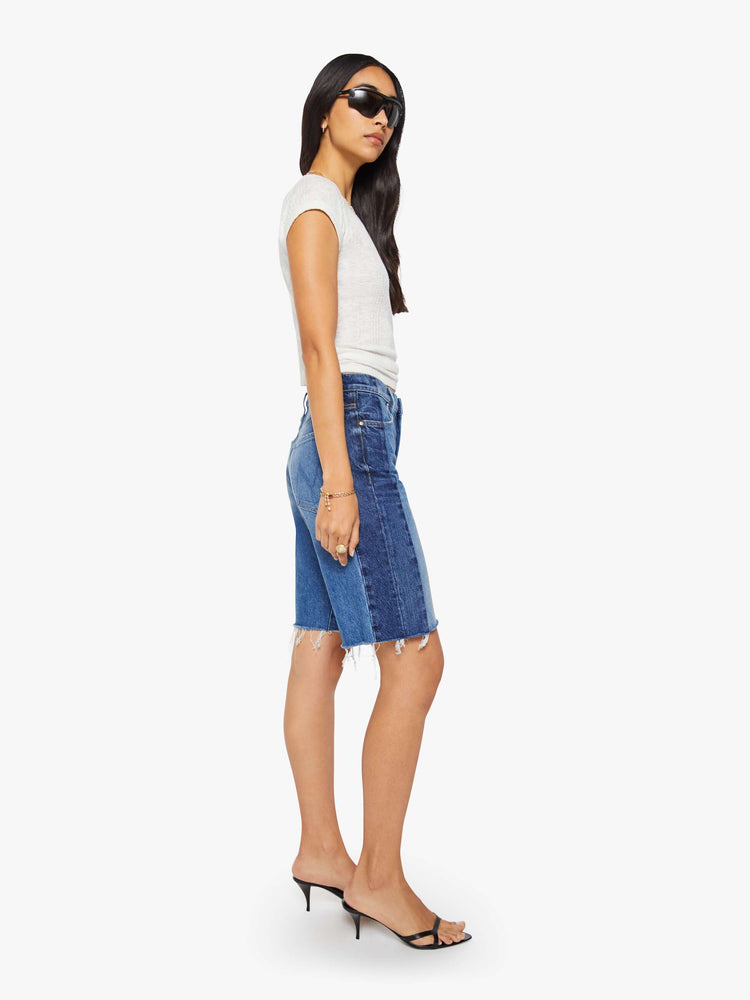 Side view of a woman in mid-blue with whiskering, fading and darker denim panels on the outer seams bermuda shorts with a mid rise, loose, wide leg, zip fly and a knee-length inseam with a frayed hem.