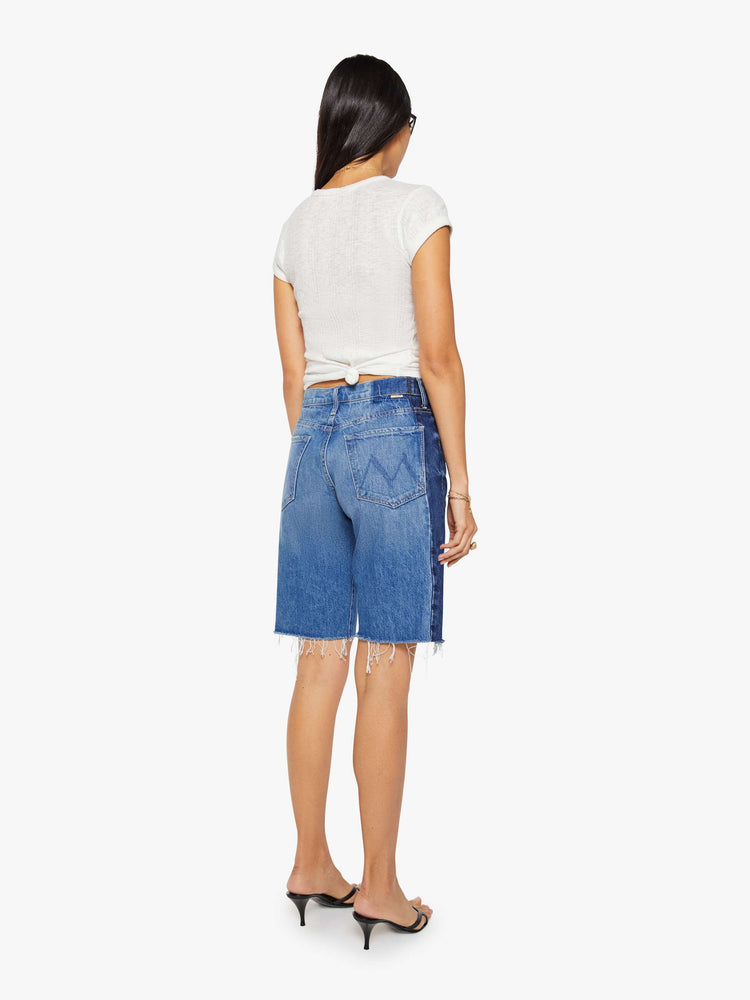 Back view of a woman in mid-blue with whiskering, fading and darker denim panels on the outer seams bermuda shorts with a mid rise, loose, wide leg, zip fly and a knee-length inseam with a frayed hem.