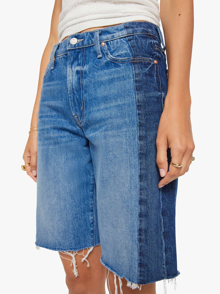 Close up view of a woman in mid-blue with whiskering, fading and darker denim panels on the outer seams bermuda shorts with a mid rise, loose, wide leg, zip fly and a knee-length inseam with a frayed hem.