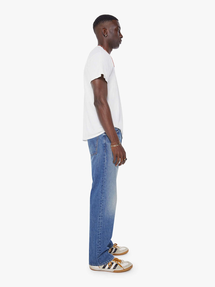 Side view of a man in a mid-blue loose-fitting jean with a mid rise, zip fly, 31-inch inseam and a clean hem.