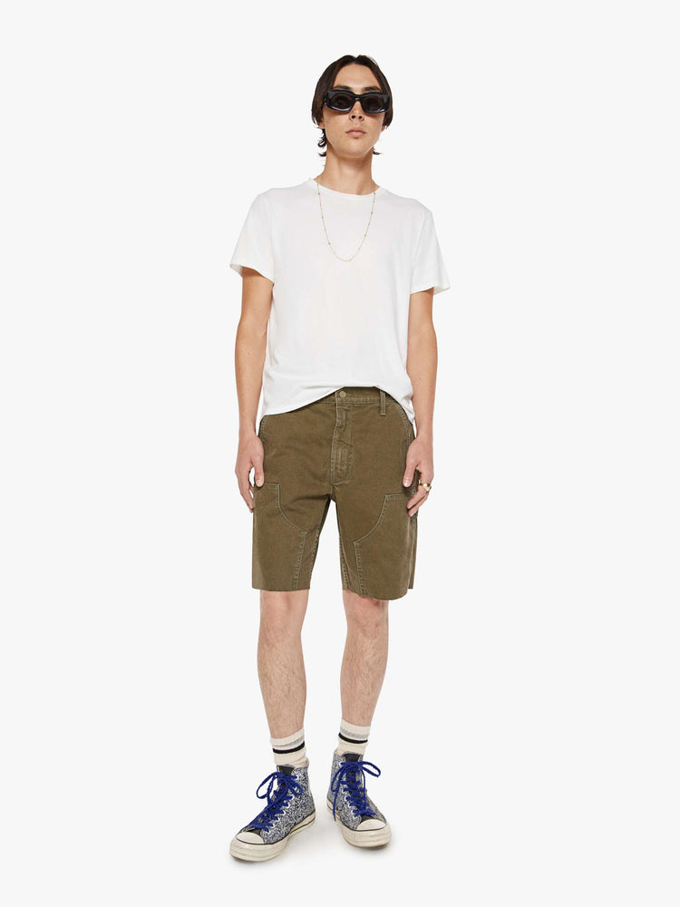 Front view of a man in a dark olive mid-rise shorts with a zip fly, slit pockets, a hammer loop, utility-inspired patches at the knees and a raw, cut-off hem.